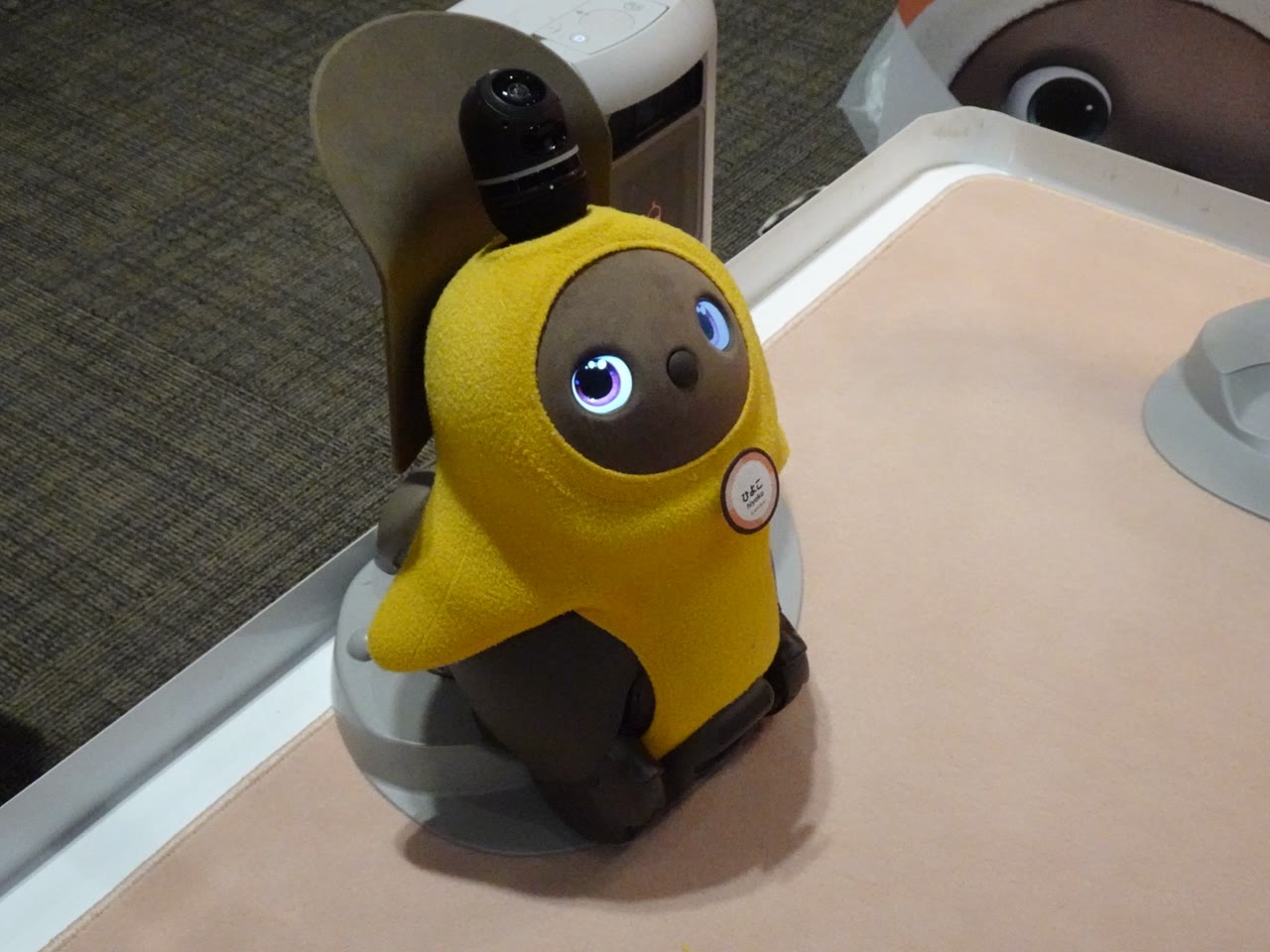A yellow-clothed earlier-generation LOVOT looking upward beside its charging nest